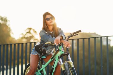 Moda kotlu çekici genç kız şık gözlük takıyor, bisiklette oturuyor ve yaz akşamları kameraya poz veriyor. Yavaş çekim