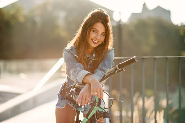 Привлекательная заманчивая девушка в джинсовой одежде носит стильные очки, сидит на велосипеде и позирует на камеру в летний вечер. Медленное движение — стоковое фото