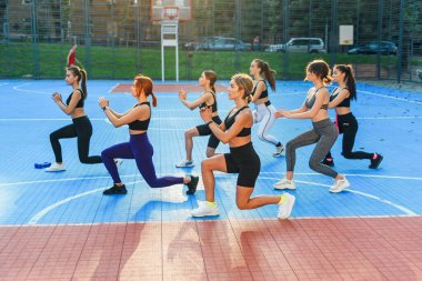 Spor giyimli çekici genç atletik kızlardan oluşan bir grup yeşil parktaki spor sahasında çömelme egzersizleri yapıyorlar.