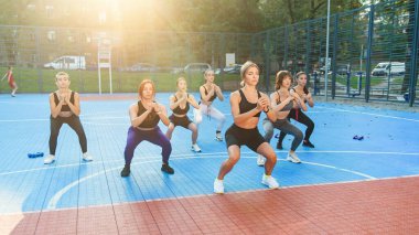Spor giyimli çekici genç atletik kızlardan oluşan bir grup yeşil parktaki spor sahasında çömelme egzersizleri yapıyorlar.