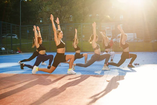 Группа привлекательных молодых атлетичных девушек в спортивной одежде делает упражнения с приседаниями на спортивной площадке в зеленом парке — стоковое фото