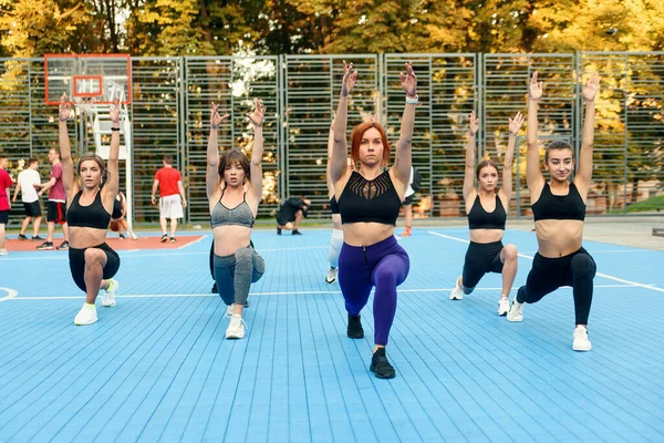 Группа привлекательных молодых атлетичных девушек в спортивной одежде делает упражнения с приседаниями на спортивной площадке в зеленом парке — стоковое фото