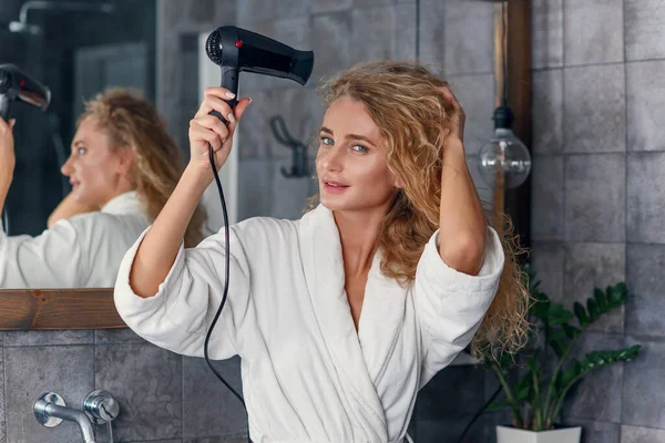 Detailní záběr půvabné usměvavé mladé dámy v bílém županu pózovat na kameru, zatímco sušení její kudrnaté vlasy pomocí vysoušeče vlasů v blízkosti koupelny zrcadlo — Stock fotografie