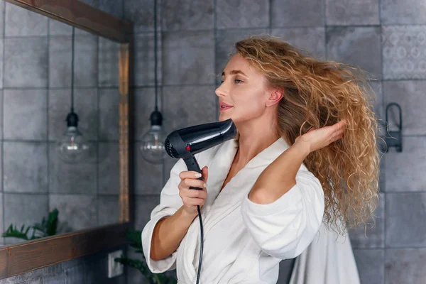 Encantadora dama sonriente con pelo rizado claro en bata blanca mirando su reflejo en el espejo del baño y secándose el cabello con un drayer — Foto de Stock