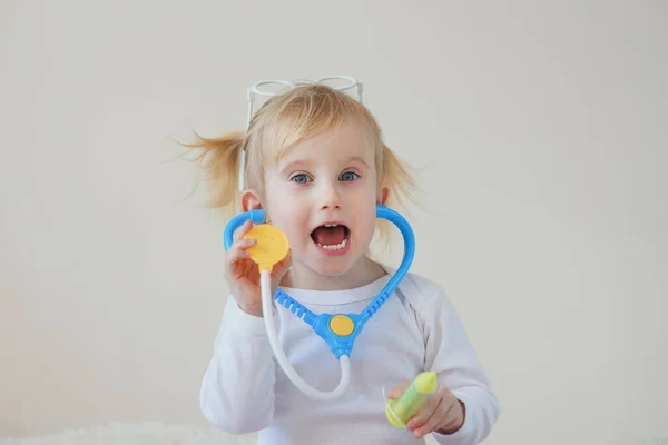 Χαρούμενο χαρούμενο κοριτσάκι φαντάζεται ως γιατρό στο νοσοκομείο χρησιμοποιώντας στηθοσκόπιο παιχνιδιών και σύριγγα. — Φωτογραφία Αρχείου