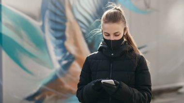 Siyah steril tıbbi maske takan genç bir kız. Kadın telefonu kullanarak NCov 2019 'la ilgili haber arıyor. Karantina COVID-19 pandemik koronavirüs salgını ve sağlık konsepti