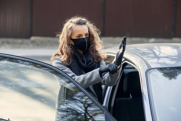 Woman in protective facial mask is wearing rubber gloves for protect herself from virus infection near car. Protective mask during quarantine, pandemic coronavirus, covid 19, contagion.