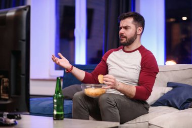 İyi görünümlü, 30 yaşında, bakımlı sakallı, evinde en sevdiği futbol takımı maçının duygusal eleştirisi sırasında çiplerin tadını çıkaran bir adam.