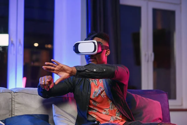 Excited young african-american man extending hand forward, using virtual reality headset sitting on bed, copy space