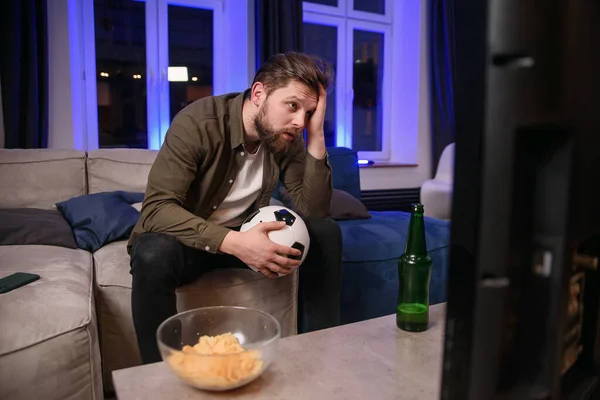 Agitated likable 30-aged bearded guy holding football ball in his hand dissatisfied from the bad game of his favourite team and falling on the couch