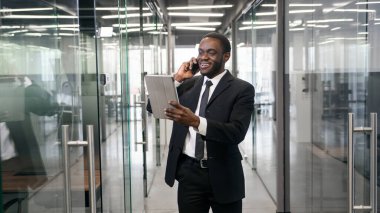 Şirket binasında yürürken ve Tablet PC 'de e-posta mesajlarını kontrol ederken akıllı telefondan konuşan Afrikalı Amerikalı iş adamı. Başarılı iş adamı konsepti.
