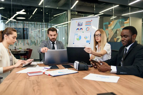 Compañeros de carreras mixtas analizando diferentes papeles de negocios y trabajando con computadoras en las mesas de la oficina moderna . — Foto de Stock