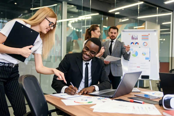 Schwarzer Büroleiter unterschreibt die Finanzunterlagen für Sekretärin in seinem Büro mit Kollegen im Hintergrund. — Stockfoto