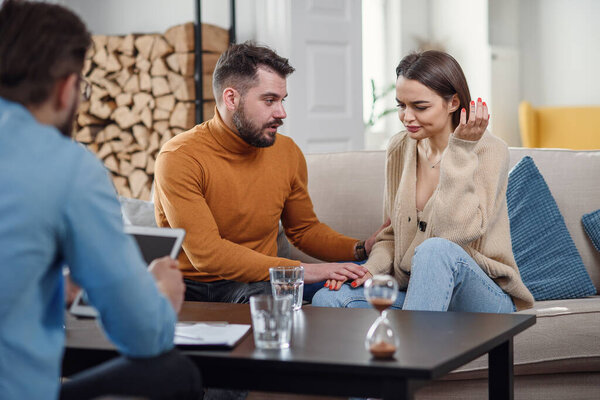 Раздраженная кавказская пара мужчин и женщин беседуют с психологом на сеансе терапии в светлой комнате
.