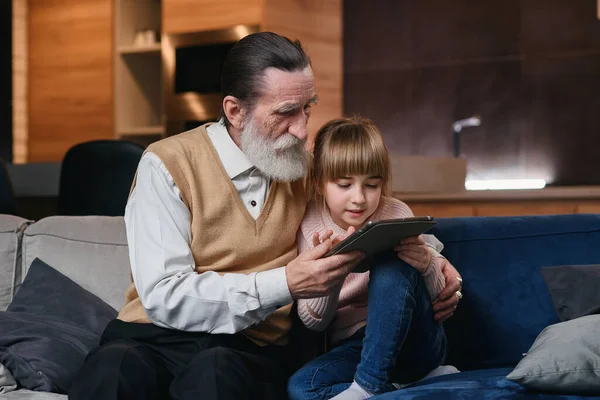 Abuelo con su nieta usando tableta PC en casa acogedora. Niña enseñarle a su abuelo a usar dispositivos inteligentes . — Foto de Stock