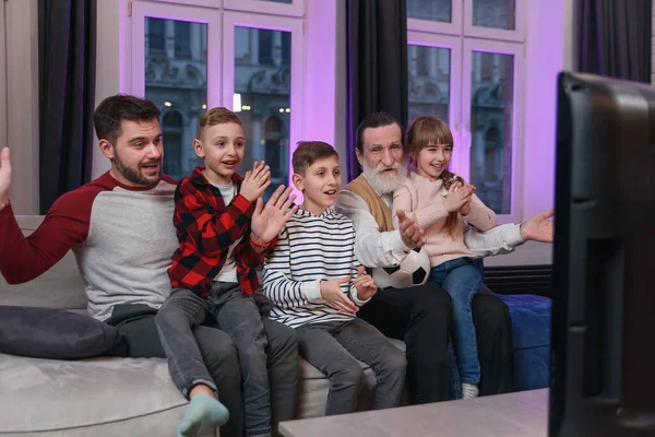 Attractive three generations of people as father, grandpa and grandchildren which sitting on the comfortable couch at home and enjoying leisure watching soccer match, shouting when team scored ball.