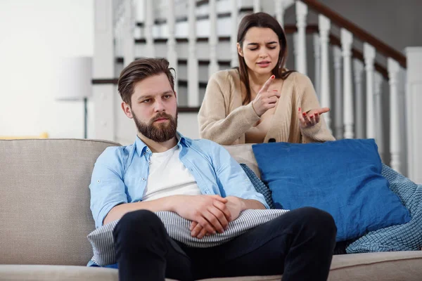 Confused man sits on the couch while his girlfriend yells and quarrels with him at home. Big family problems. — Stock Photo, Image