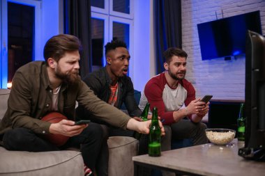 Takım oyunlarından memnun olmayan uluslararası erkek arkadaşlar bira içip akıllı telefonlarını kurcalayarak TV 'de dünya şampiyonasını izliyorlar..