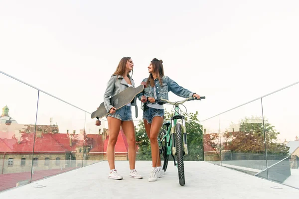 Retrato de duas jovens mulheres urbanas atraentes andando com uma bicicleta e skate na rua no dia ensolarado de verão. Movimento lento . — Fotografia de Stock