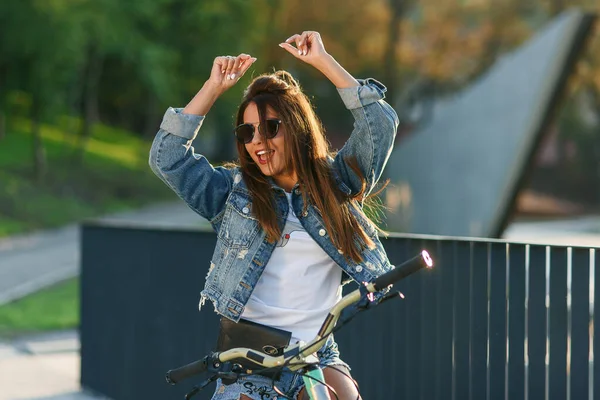 Atraktivní lákavé mladé dívky v módních džínách oblečení nosí stylové brýle, sedí na kole a pózuje na kameru v létě večer. Zpomalený pohyb — Stock fotografie