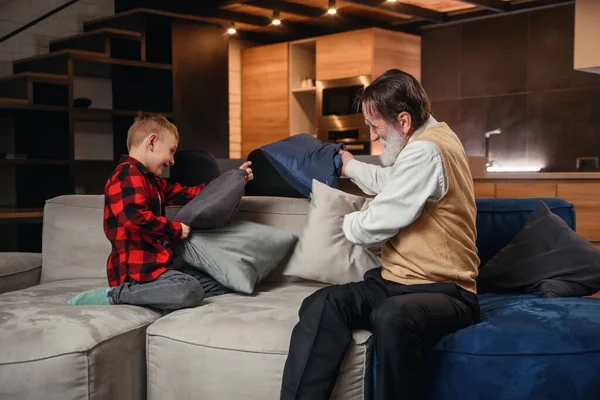 Happy boy with funny modern grandpa fighting with pillows having fun together sitting on comfortable couch at home.