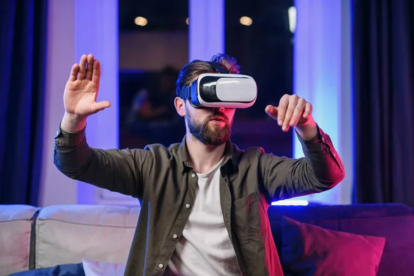 Attractive bearded man enjoying virtual reality glasses home in the evening. Smartphone use with VR goggles headset.