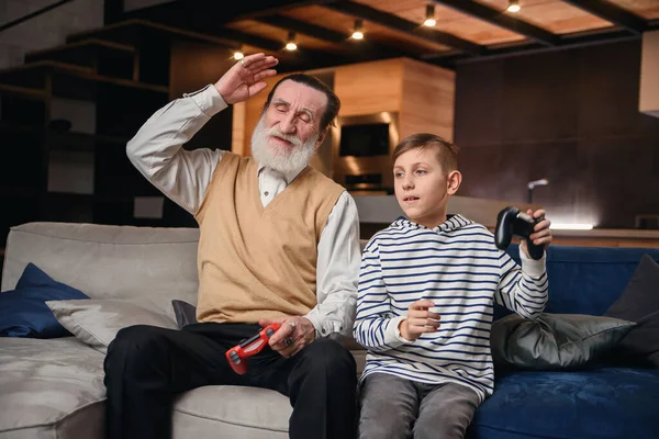 Menino bonito com o avô sentado no sofá e jogando videogame com almofada de jogo — Fotografia de Stock