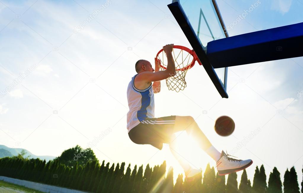 Young basketball player drives to the hoop for a high flying slam dunk in front of sunset sky.