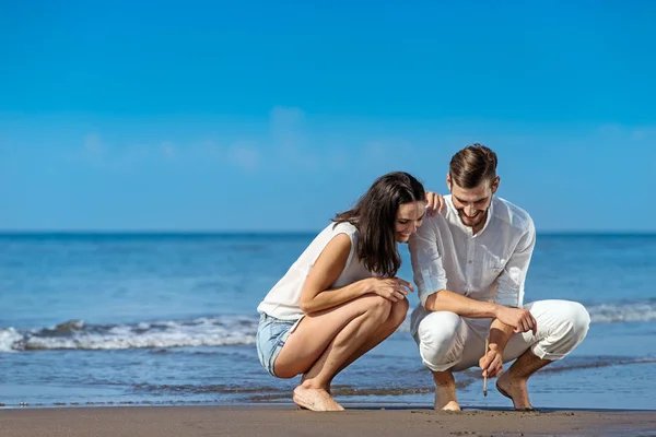 Romantische jong koppel tekenen vormen op het zand terwijl op huwelijksreis. — Stockfoto