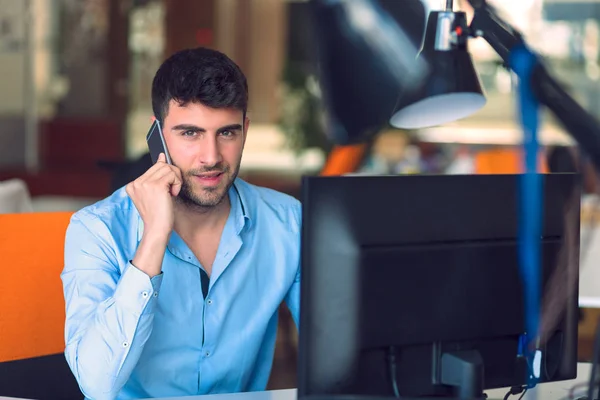 Jovem empresário ocupado trabalhando no computador portátil enquanto fala no smartphone no escritório . — Fotografia de Stock