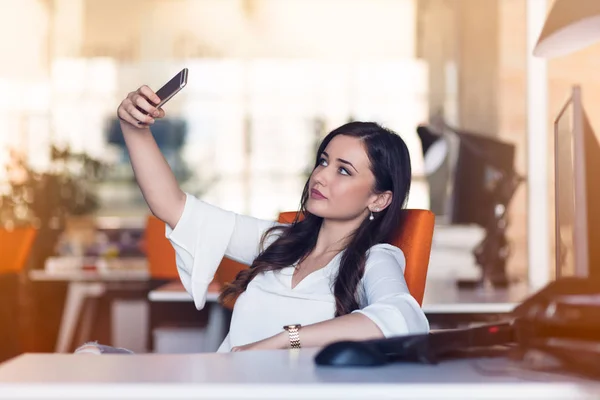 Krásná mladá žena dělá selfie v její pracoviště. Ona uvolňuje a dát nohy na stůl. — Stock fotografie