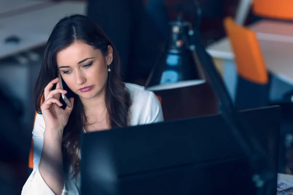 Beautiful young woman working on computer and talking on phone — Stock Photo, Image
