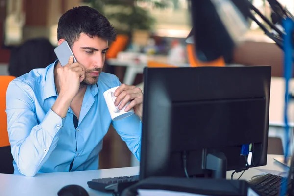 Jovem empresário ocupado trabalhando no computador portátil enquanto fala no smartphone no escritório . — Fotografia de Stock