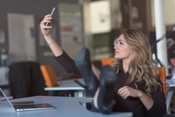 Krásná mladá žena dělá selfie v její pracoviště. Ona uvolňuje a dát nohy na stůl. — Stock fotografie