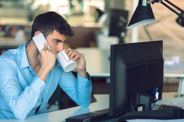 Jovem empresário ocupado trabalhando no computador portátil enquanto fala no smartphone no escritório . — Fotografia de Stock