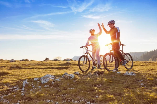 Feliz pareja va en una montaña asfalto carretera en el bosque en bicicletas con cascos dando uno al otro un alto cinco —  Fotos de Stock