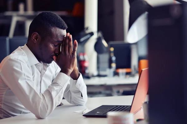 Втомився африканських людини, що сидить в офісі після жорсткий робочий день, працює на ноутбуці, намагаючись зосередитися — стокове фото