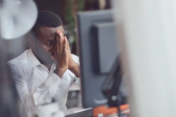 Κουρασμένος Αφρικής άνδρας κάθεται σε ένα γραφείο μετά από ένα σκληρό εργάσιμης ημέρας, λειτουργεί σε φορητό υπολογιστή, προσπαθεί να συγκεντρώσει — Φωτογραφία Αρχείου