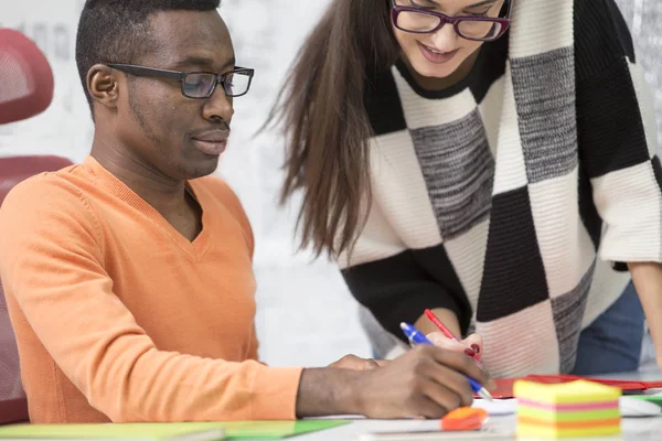 Twee uiteenlopende werken collega's glimlachend en het neerschrijven van notities samen zittend aan een tafel in een modern kantoor — Stockfoto