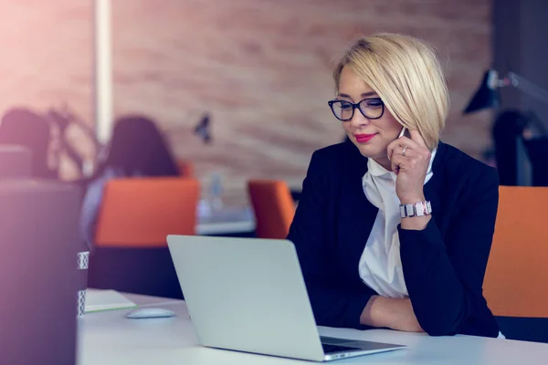 Mulher de negócios loira encantadora atraente falando em um celular enquanto está sentado na mesa de escritório trabalhando em um laptop . — Fotografia de Stock
