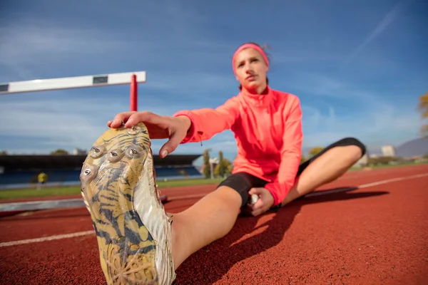 Young sportswoman sitting and stretching on track field while listening to music, looking at camera. — Stock Photo, Image