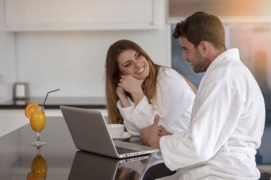 Genç çift bornoz evde mutfakta kahve içer ve dizüstü bilgisayar üzerinde çalışan