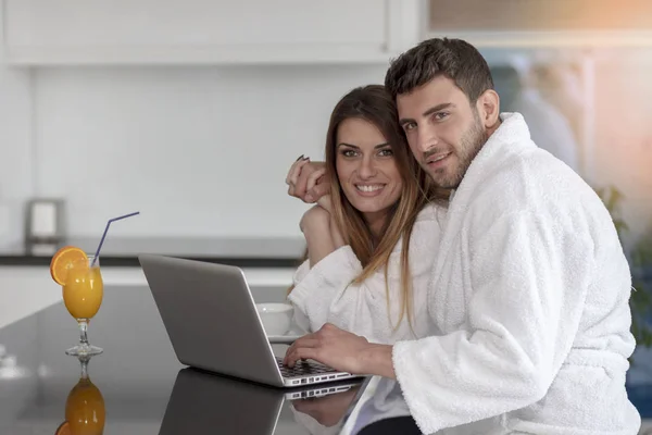 Pareja joven en albornoz en casa tomando café en la cocina y trabajando en el ordenador portátil — Foto de Stock