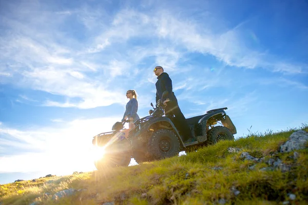 Hermosa pareja está viendo la puesta de sol desde la montaña sentado en quadbike atv —  Fotos de Stock