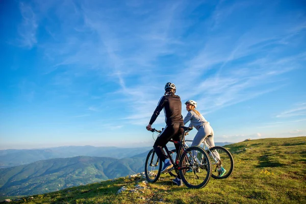 Cyclist couple with mountain bikes standing on the hill under the evening sky and enjoying bright sun at the sunset. — Stock Photo, Image