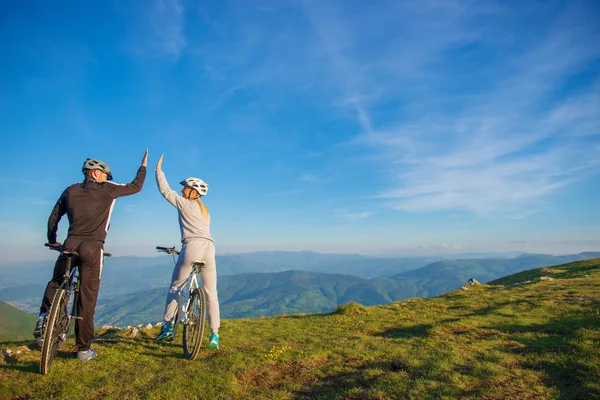 Feliz pareja va en un camino de montaña en el bosque en bicicletas con cascos dando unos a otros un alto cinco —  Fotos de Stock