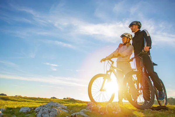 Pareja ciclista con bicicletas de montaña de pie en la colina bajo el cielo de la noche y disfrutando del sol brillante al atardecer . —  Fotos de Stock