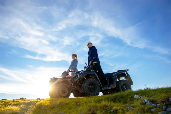 Hermosa pareja está viendo la puesta de sol desde la montaña sentado en quadbike atv —  Fotos de Stock