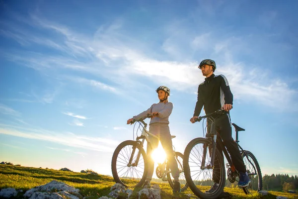 Pareja ciclista con bicicletas de montaña de pie en la colina bajo el cielo de la noche y disfrutando del sol brillante al atardecer . —  Fotos de Stock