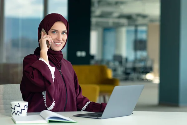 Heureuse femme d'affaires musulmane en hijab au bureau. Femme arabe souriante travaillant sur un ordinateur portable et parlant sur un smartphone — Photo